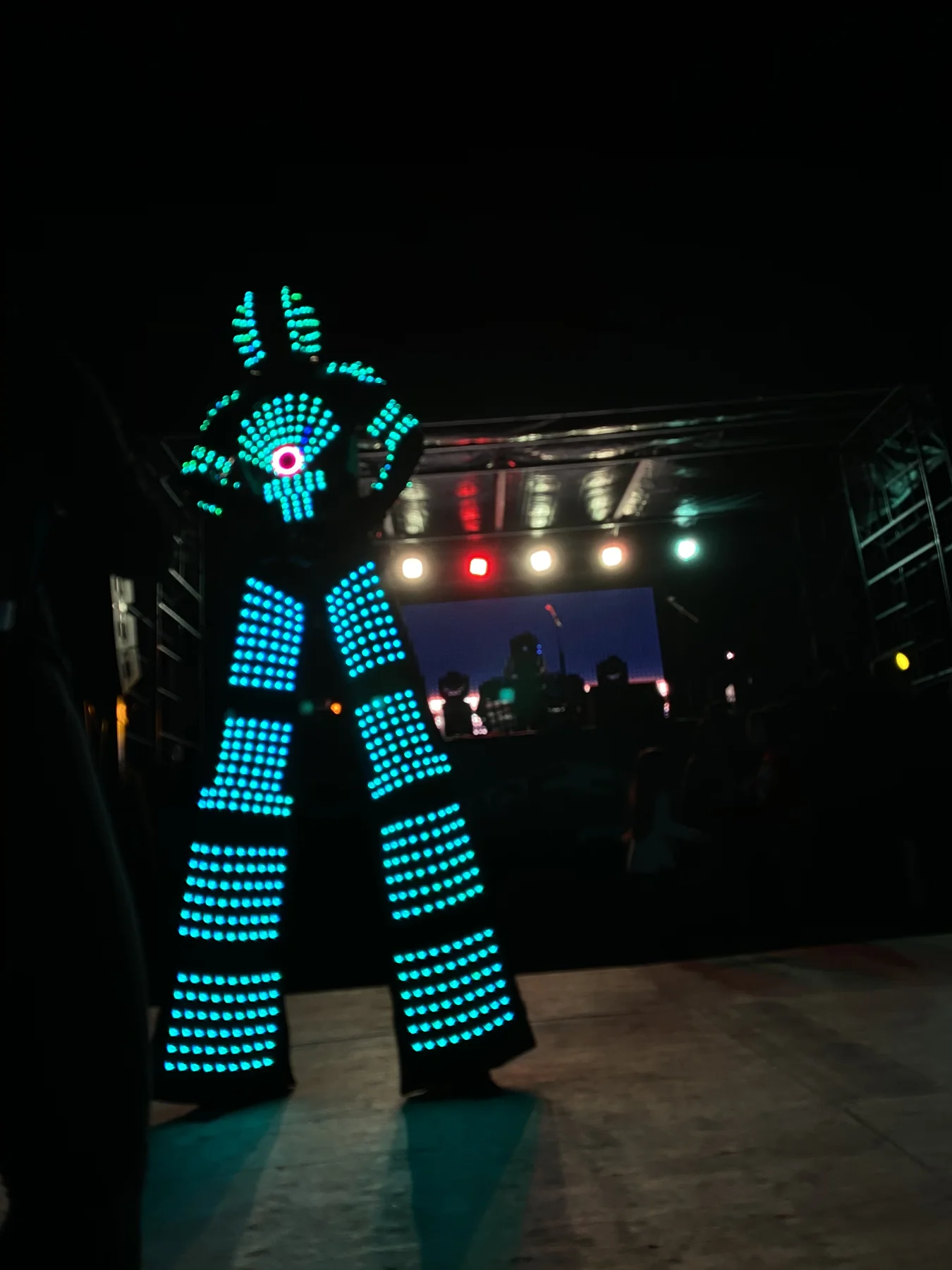 An illuminated LED robot performer near an outdoor stage